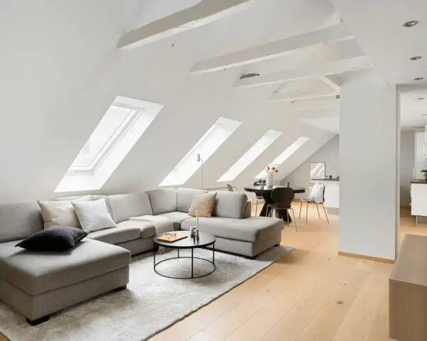 Soggiorno mansardato con divano angolare grigio, tavolino nero e travi a vista moderne dipinte di bianco che creano un'atmosfera luminosa.