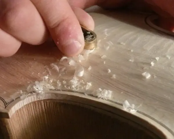 Hände bearbeiten mit einem Werkzeug das helle Holz einer Geige, Späne fliegen. Die Frage "aus welchem Holz besteht die Geige" wird hier visuell beantwortet.