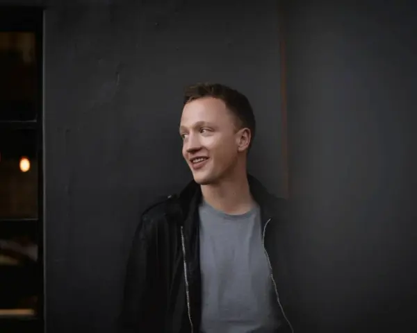 Joris, lächelnd, lehnt an einer dunklen Wand.