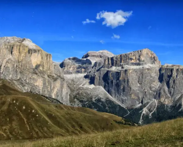 Vaste cime rocciose del gruppo Sella Dolomiti sotto un cielo azzurro con nuvole bianche. Prati verdi e sentieri serpeggiano sui pendii.