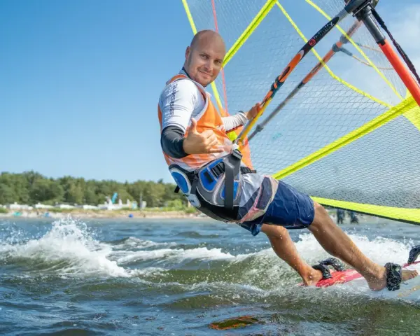 Mężczyzna na desce windsurfingowej, uśmiecha się i pokazuje kciuk w górę.
