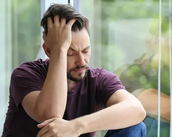 Mann sitzt mit geschlossenen Augen und der Hand am Kopf vor einem Fenster.