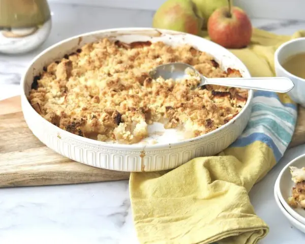 Ein goldbrauner Apfel-Crumble in einer Auflaufform, bereit zum Servieren. Daneben eine Tasse Tee und frische Äpfel.