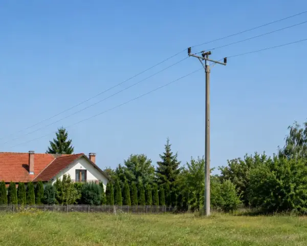 Słup na działce, obok domu z czerwoną dachówką i żywopłotem. W tle zielone drzewa i błękitne niebo.