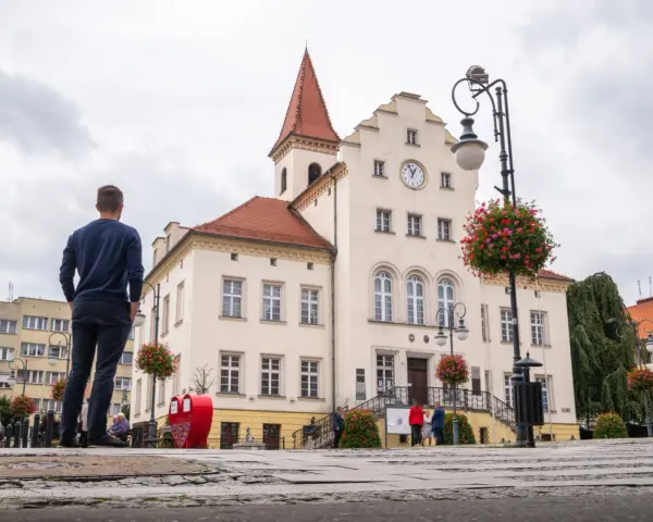 Mężczyzna podziwia ratusz, jedną z głównych trzebnickich atrakcji. W tle kwitnące pelargonie i zabytkowa architektura.