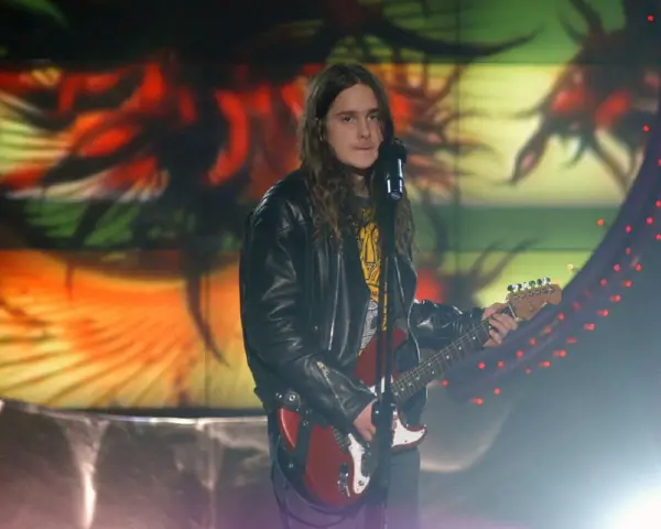 Zalewski w Idolu, z gitarą i w skórzanej kurtce, na tle kolorowej scenografii.