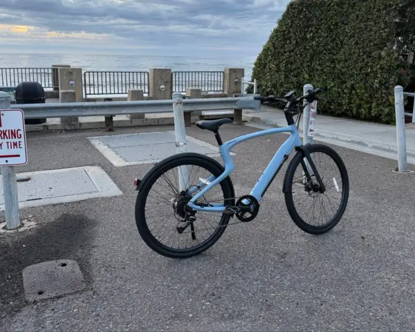 A light blue electric city bike is parked on asphalt near a "No Parking" sign, with the ocean and cloudy sky in the background.
