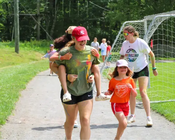 Mädchen haben Spaß bei einem Sommercamp in den USA. Eine trägt die andere auf dem Rücken, während sie lachend über einen Weg laufen.
