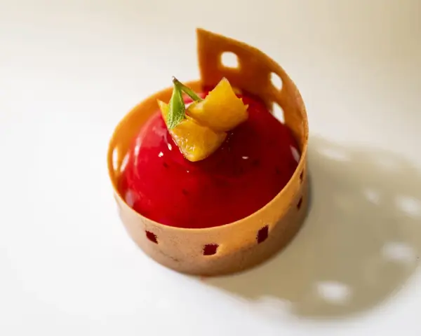 A red berry mousse dessert with orange zest and mint, presented in a decorative pastry shell on a white plate.