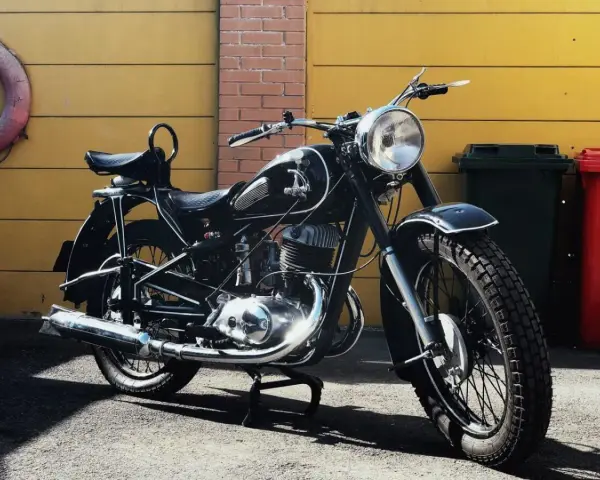 Klasyczny czarny motocykl, model iż 49, lśni w słońcu. Obok żółta brama i ceglana ściana.