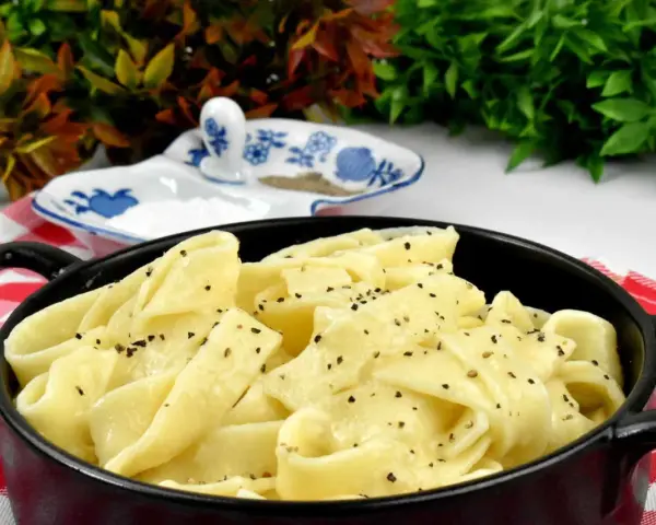 Pappardelle w kremowym sosie alfredo, posypane świeżo mielonym pieprzem, podane w czarnym naczyniu na czerwono-białej kratce.
