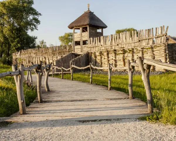 Drewniana kładka prowadzi do grodziska, jednego z historycznych miejsc w Polsce. Widać palisadę i wieżę obronną.