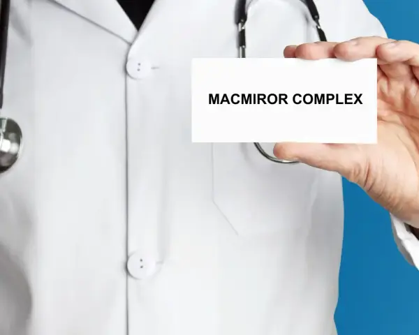 Lekarz w białym kitlu prezentuje kartkę z napisem MACMIROR COMPLEX. Stetoskop wisi na jego szyi.