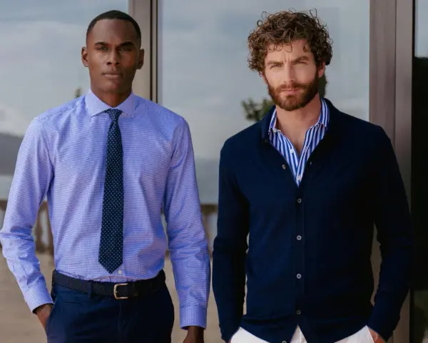 Dos hombres posan. Uno lleva una camisa de vestir azul con corbata, la camisa por dentro. El otro, una camisa de rayas azules y blancas con cárdigan azul oscuro, la camisa por fuera.