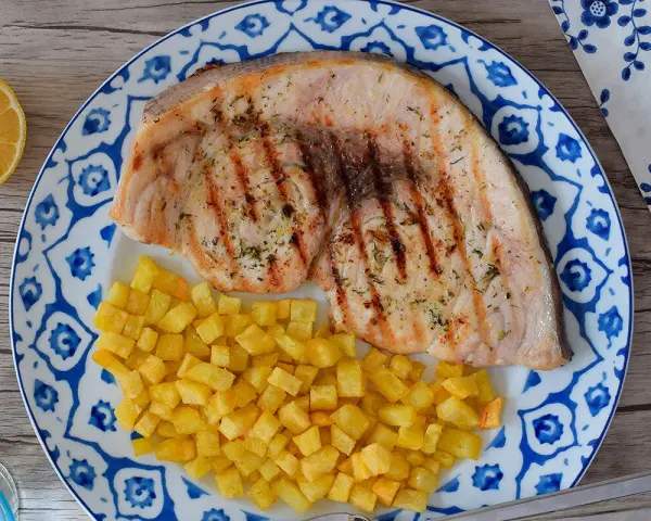 Delicioso emperador a la plancha con patatas fritas crujientes, servido en un plato con diseño azul y blanco. Un toque de limón completa esta apetitosa presentación.