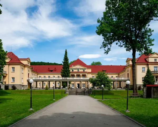 Eleganckie, żółte budynki z czerwonymi dachami, przypominające stare łazienki Krynica Zdrój, otoczone zielonym ogrodem.