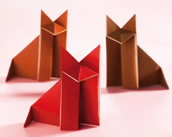 Drei origami Füchse, einer rot, zwei braun, sitzen auf rosa Untergrund.