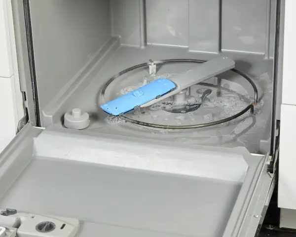 Water and suds fill the bottom of a dishwasher, with a blue spray arm component visible. This image shows a step in how to fix a clogged dishwasher.