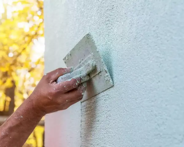 Dłoń zacierająca tynk, kontrolując jego grubość. W tle jesienne drzewa.