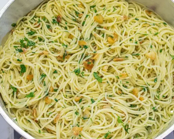 Espaguetis al ajillo recién hechos, con trocitos de ajo dorado y perejil fresco, servidos en una sartén metálica.