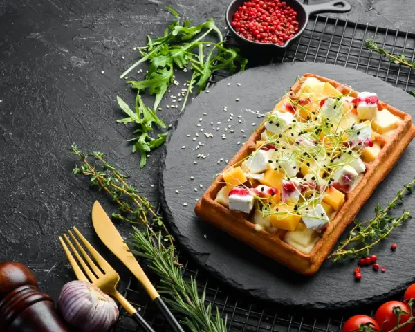 Gaufre rectangulaire garnie de dés de fromage, de fruits et de pousses, servie sur une ardoise. Une délicieuse pâte à gaufre salée prête à être dégustée.