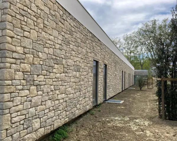 Un mur extérieur en pierres naturelles irrégulières, avec des fenêtres modernes et un toit blanc. Le sol est en terre battue.