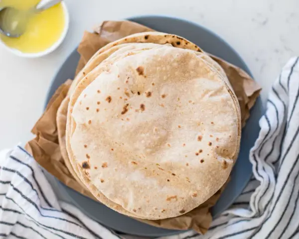 Stos świeżo upieczonych, lekko przypieczonych roti na talerzu, obok miseczki z roztopionym masłem.