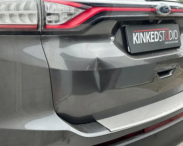 Widoczne wgniecenie w tylnej klapie szarego Forda. Tabliczka "KINKEDSTUDIO" sugeruje miejsce naprawy.