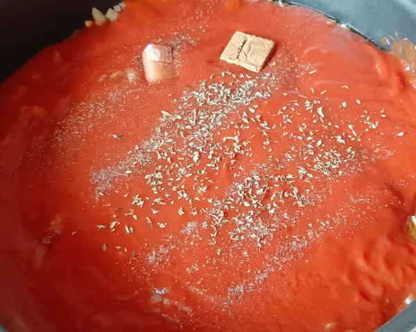 Un coulis de tomates mijote dans une poêle, agrémenté d'herbes et d'un cube de bouillon.