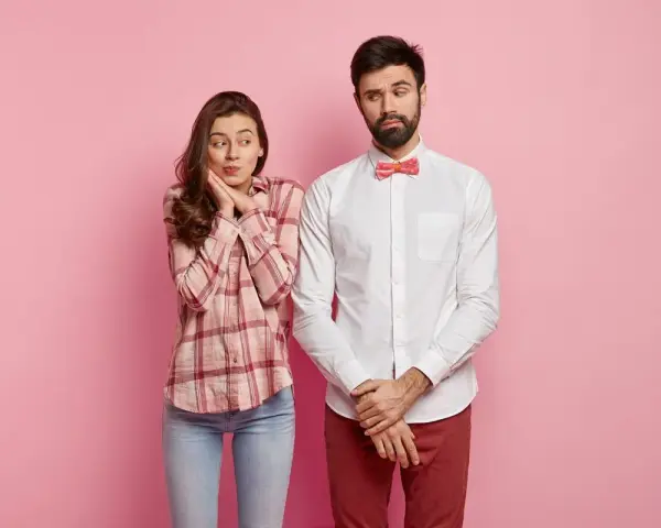 Coppia su sfondo rosa. Lei, con camicia a quadri e jeans, sembra timida. Lui, con camicia bianca e papillon, è pensieroso. Come vestirsi al primo appuntamento uomo?