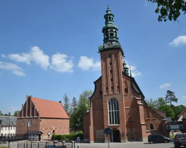 Majestatyczna kolegiata w Kartuzach z cegły, z zieloną wieżą i zegarem, na tle błękitnego nieba z chmurami.