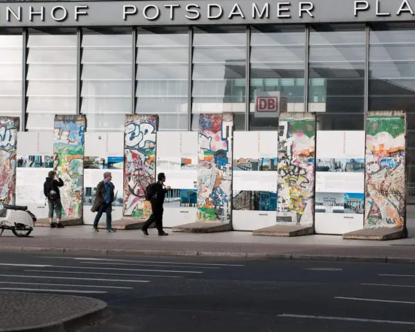 Bunte Reste des Berliner Mauer-Denkmals vor dem Bahnhof Potsdamer Platz. Menschen spazieren vorbei, ein Roller parkt daneben.