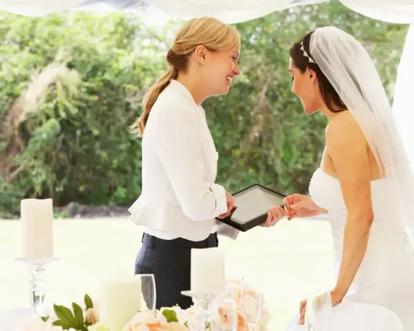 Panna młoda z welonem i wedding plannerka dyskutują przy stole. Czy warto wynająć wedding plannera, czy poradzić sobie samemu?