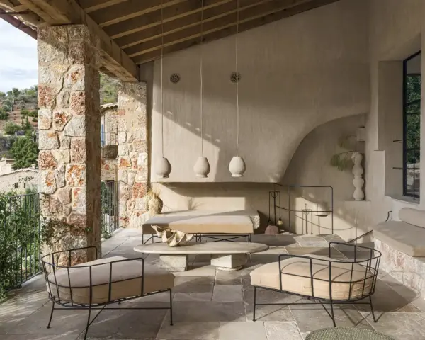 Acogedora terraza interior piso con sillones de metal, sofá y mesa de centro de piedra, bajo un techo de madera y rodeada de muros de piedra.