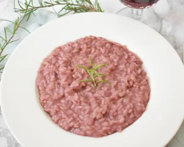 Różowe risotto z prawdziwkami i rozmarynem na białym talerzu, obok widelca i kieliszka wina.