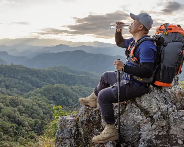 Backpacker drinks water on a rocky outcrop, contemplating how much water to bring backpacking for this stunning mountain vista.