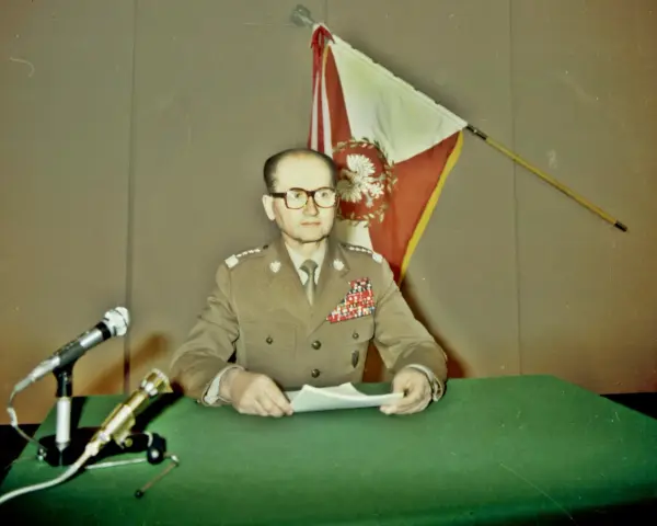 Generał w mundurze, z flagą Polski za plecami, przemawia na konferencji prasowej. To ważny moment dla Wojskowej Rady Ocalenia Narodowego.