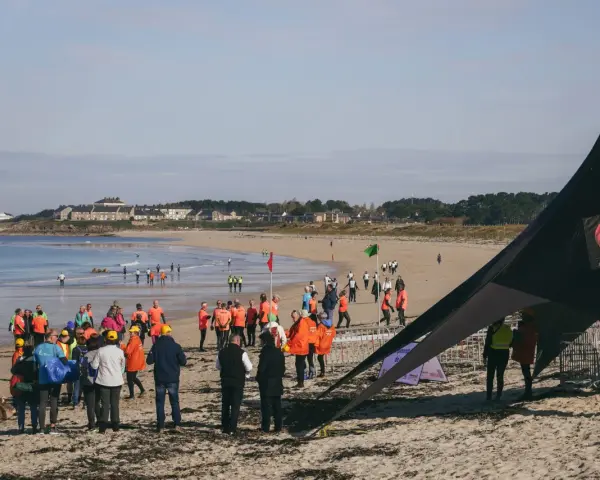 Rassemblés sur la plage pour une course de longe côte, des participants en tenues colorées attendent le départ sous un ciel clair.