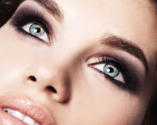 Occhi azzurri intensificati da un trucco smokey eyes audace, con labbra lucide e un accenno di sorriso.