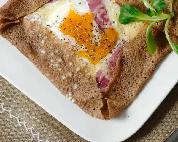 Une galette bretonne garnie d'un œuf, de jambon et de fromage, poivrée et décorée d'une feuille verte. Un délice de french crepes !