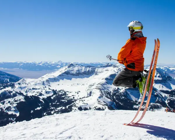 Un skieur en l'air, prêt à dévaler la pente. Il porte un casque et une veste orange vif, rappelant les règles pour skier en toute sécurité. Le paysage montagneux enneigé est spectaculaire.