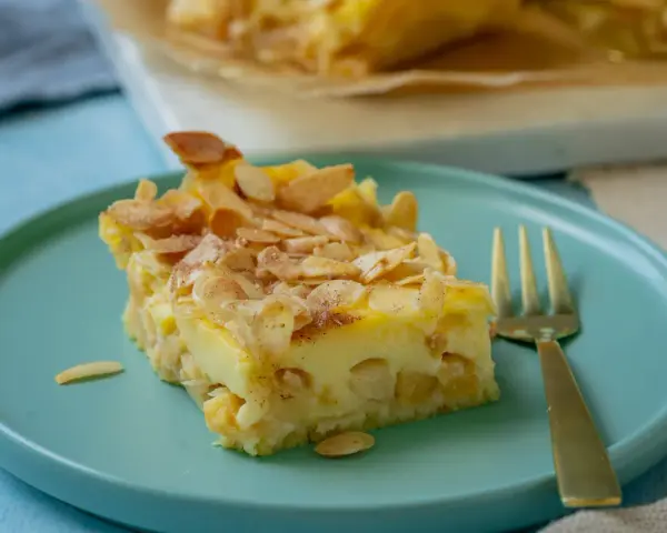 Ein Stück schneller Apfelkuchen mit Schmand und Pudding, bestreut mit Mandelblättchen und Zimt, auf einem türkisfarbenen Teller.