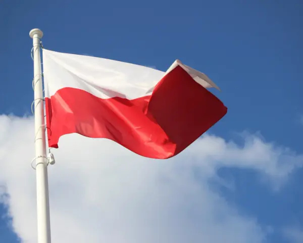 Biało-czerwona bandera polska powiewa na maszcie na tle błękitnego nieba z chmurami.