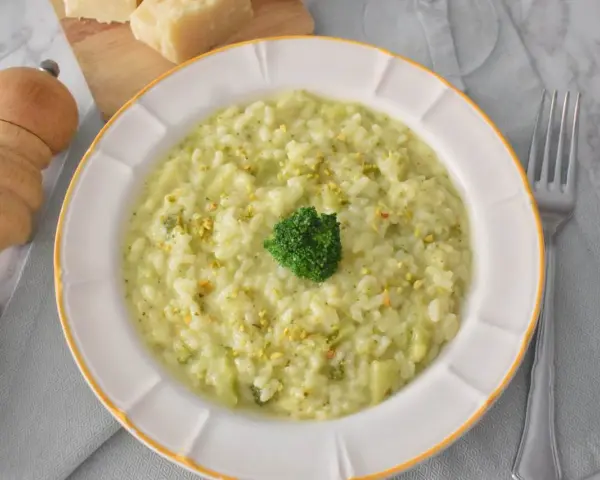 Kremowe risotto z brokułami, posypane pistacjami, z małą różyczką brokułu na wierzchu. Obok widelca i kawałków parmezanu.
