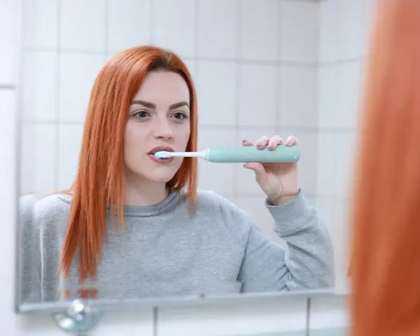 Rudowłosa kobieta myje zęby szczoteczką soniczną, zastanawiając się, czy szczoteczka soniczna usuwa kamień.