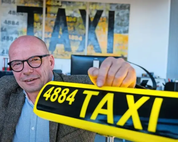 Mann mit Brille hält ein gelbes Taxi-Schild mit der Nummer 4884. Im Hintergrund Kunstwerke mit "TAXI" und der Zahl 1884.