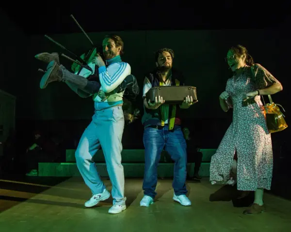 Iwo Orłowski życiorys: scena teatralna z mężczyzną niosącym krzesło i walizkę, obok kobieta w sukience.