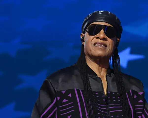 Stevie Wonder heute, mit Sonnenbrille und schwarzer Kappe, trägt eine Jacke mit lila Muster vor blauem Sternenhintergrund.