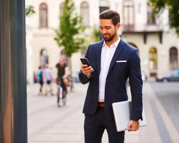 Mężczyzna w eleganckim stroju biznesowym męskim sprawdza telefon, trzymając laptopa.