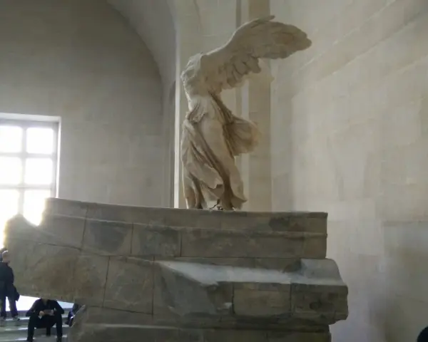 Skrzydlata Nike, symbol zwycięstwa, niczym mit, unosi się nad kamiennym postumentem.